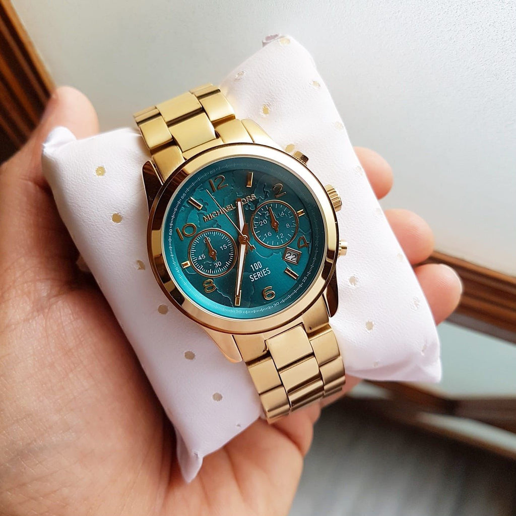 Michael Kors MK5815 gold-tone watch with turquoise world-map dial on a white pillow