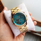 Michael Kors MK5815 gold-tone watch with turquoise world-map dial on a white pillow